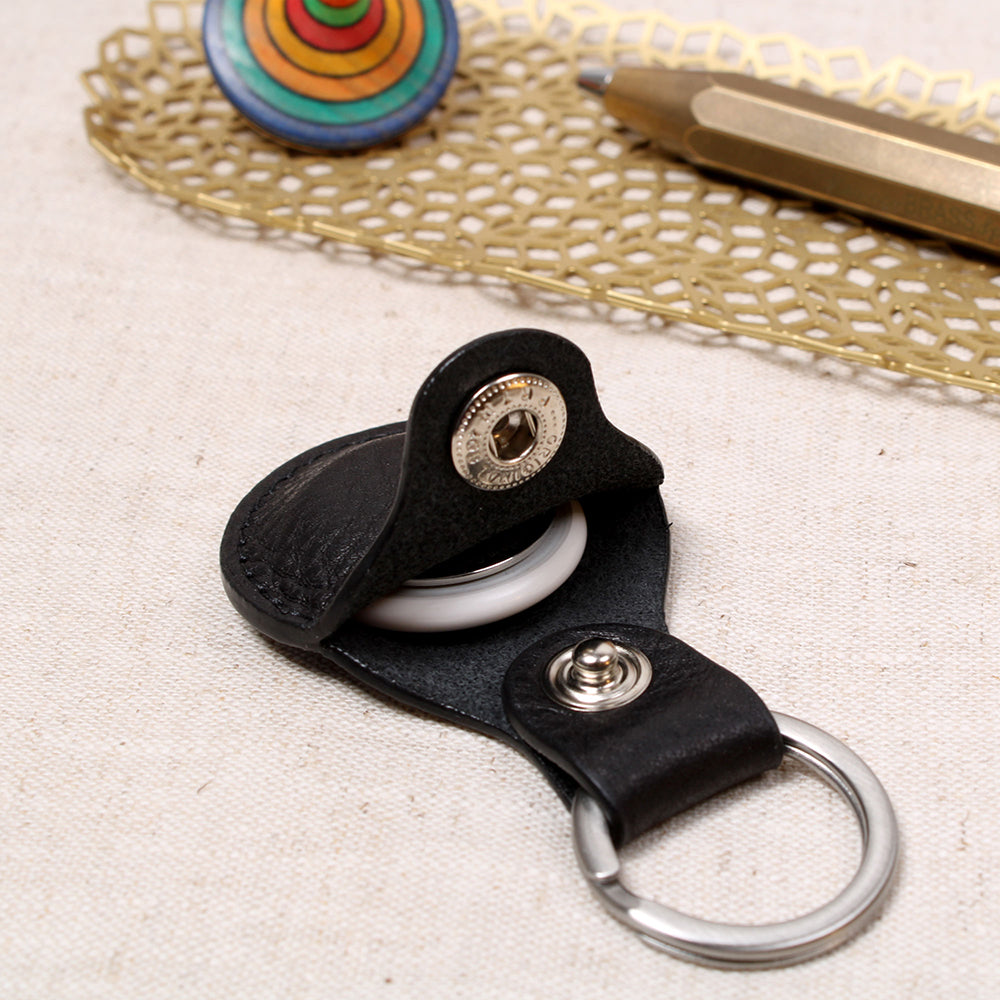 Offener AirTag Anhänger aus pflanzlich gegerbtem Leder in Schwarz von Good Wilhelm mit eingelegtem Apple AirTag, Edelstahl-Schlüsselring, liegend auf einem Holztisch