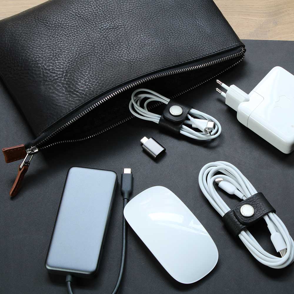 Auf einem Tisch liegt eine Kabeltasche aus Leder in Schwarz und davor liegen Kabel, Maus und Power ein Adapter