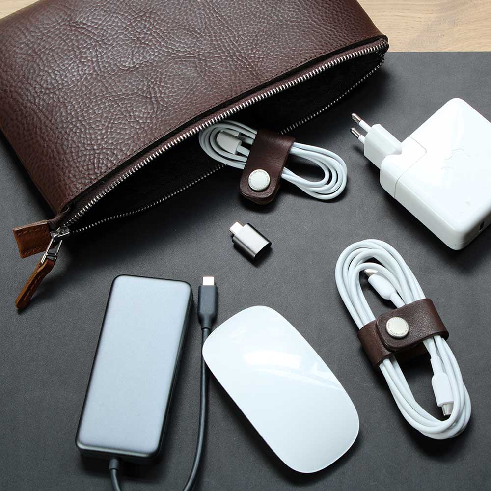 Auf einem Tisch liegt eine Kabeltasche aus Leder in Dunkelbraun und davor liegen Kabel, Maus und Power ein Adapter